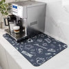 A rectangular dark gray coffee-themed mat with white chalk-style drawings and text, placed under a stainless steel espresso machine on a white marble countertop.