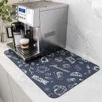 A rectangular dark gray coffee-themed mat with white chalk-style drawings and text, placed under a stainless steel espresso machine on a white marble countertop.