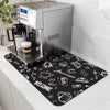 A rectangular black coffee mat with white hand-drawn style coffee-themed illustrations and words like 'WELCOME' and 'Fresh', placed on a white countertop under a stainless steel espresso machine. A coffee cup sits on the drip tray.