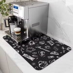 A rectangular black coffee mat with white hand-drawn style coffee-themed illustrations and words like 'WELCOME' and 'Fresh', placed on a white countertop under a stainless steel espresso machine. A coffee cup sits on the drip tray.