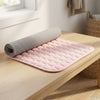 A pink quilted yoga mat with a gray textured underside, partially unrolled on a light wood table in a bright, modern room.