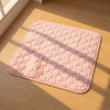 A square pink quilted cushion pad with a raised circular pattern, placed on a light wood floor in warm sunlight with soft shadows.